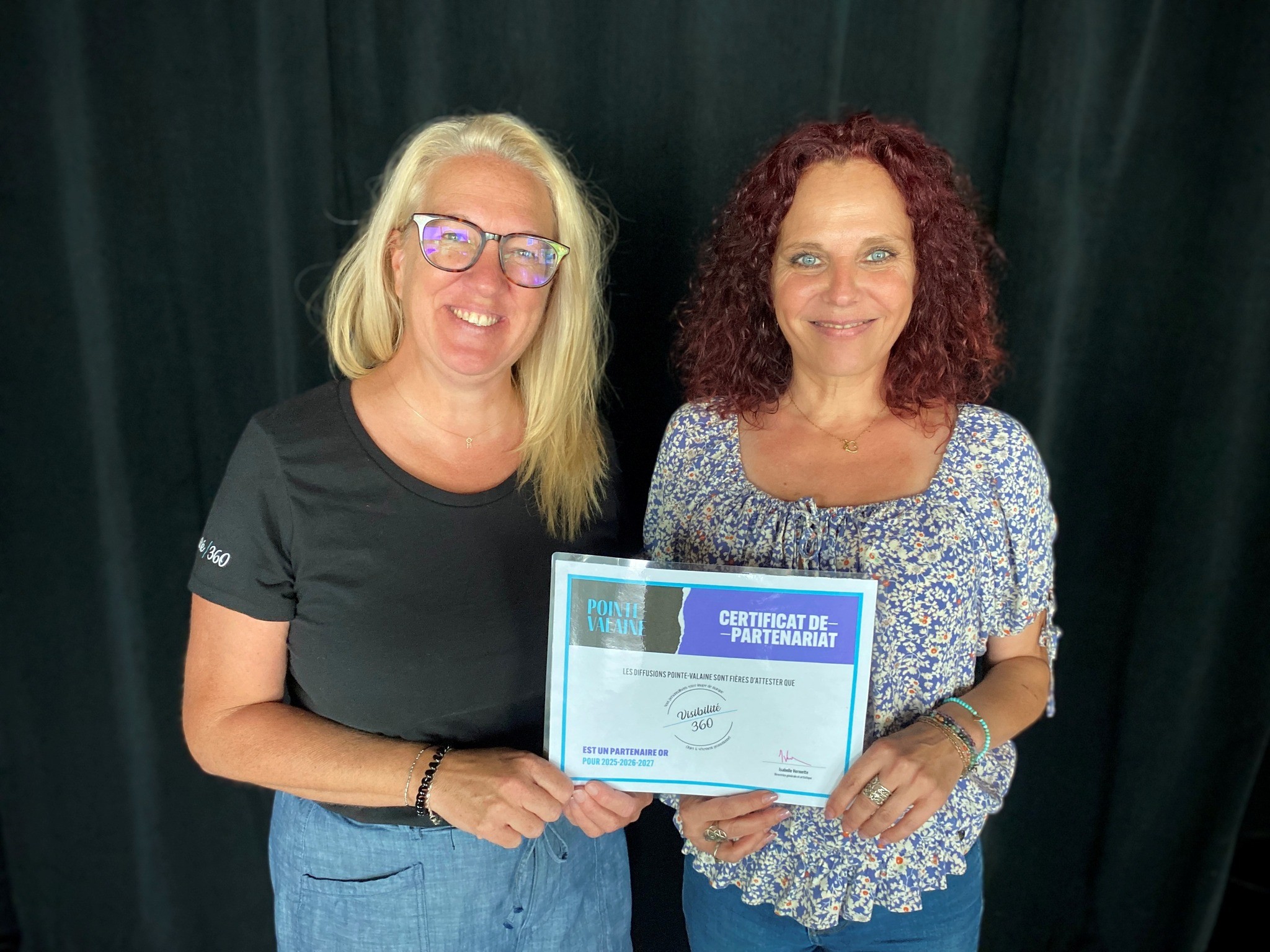Deux femmes sont debout et sourient à l'appareil photo. L'une d'elles tient un certificat avec un texte en français, et toutes deux se trouvent devant un fond sombre et uni.