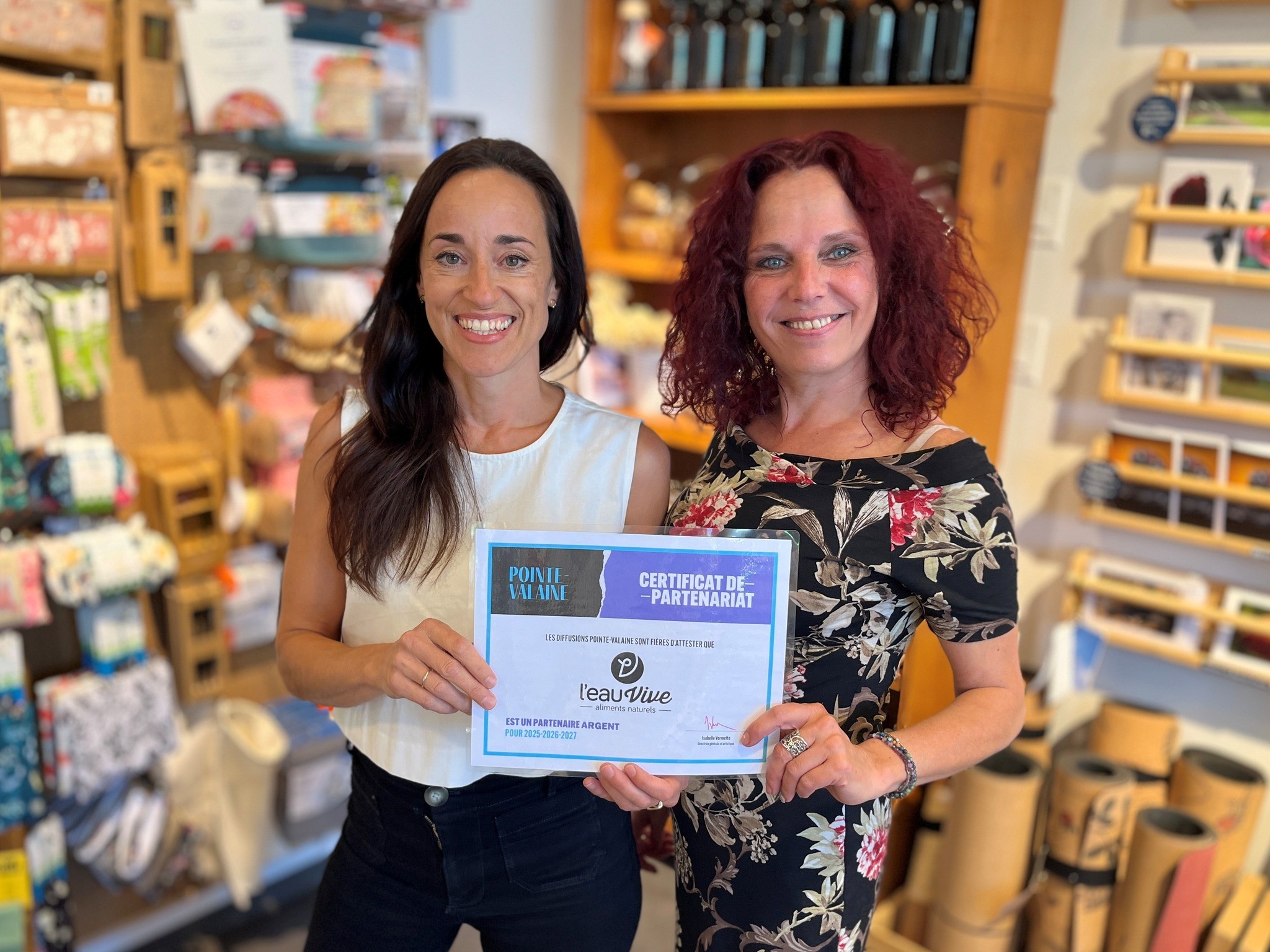 Deux femmes souriantes se tiennent à l'intérieur et tiennent un certificat de partenariat. L'arrière-plan présente des étagères avec divers produits colorés et des articles cadeaux. Les deux femmes sont habillées de façon décontractée et semblent heureuses et fières.