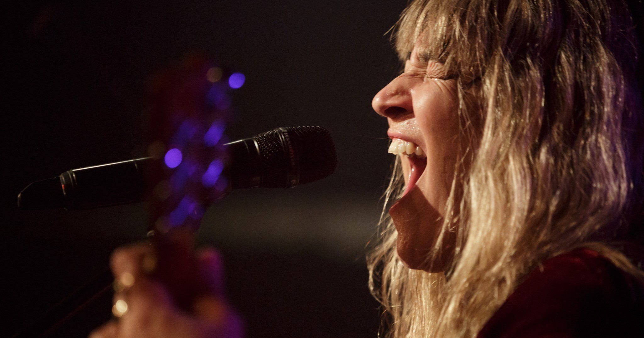 Une musicienne blonde chante passionnément dans un micro sur scène, les yeux fermés et la bouche grande ouverte, tenant une guitare, avec un éclairage dramatique mettant en valeur son visage et ses cheveux.