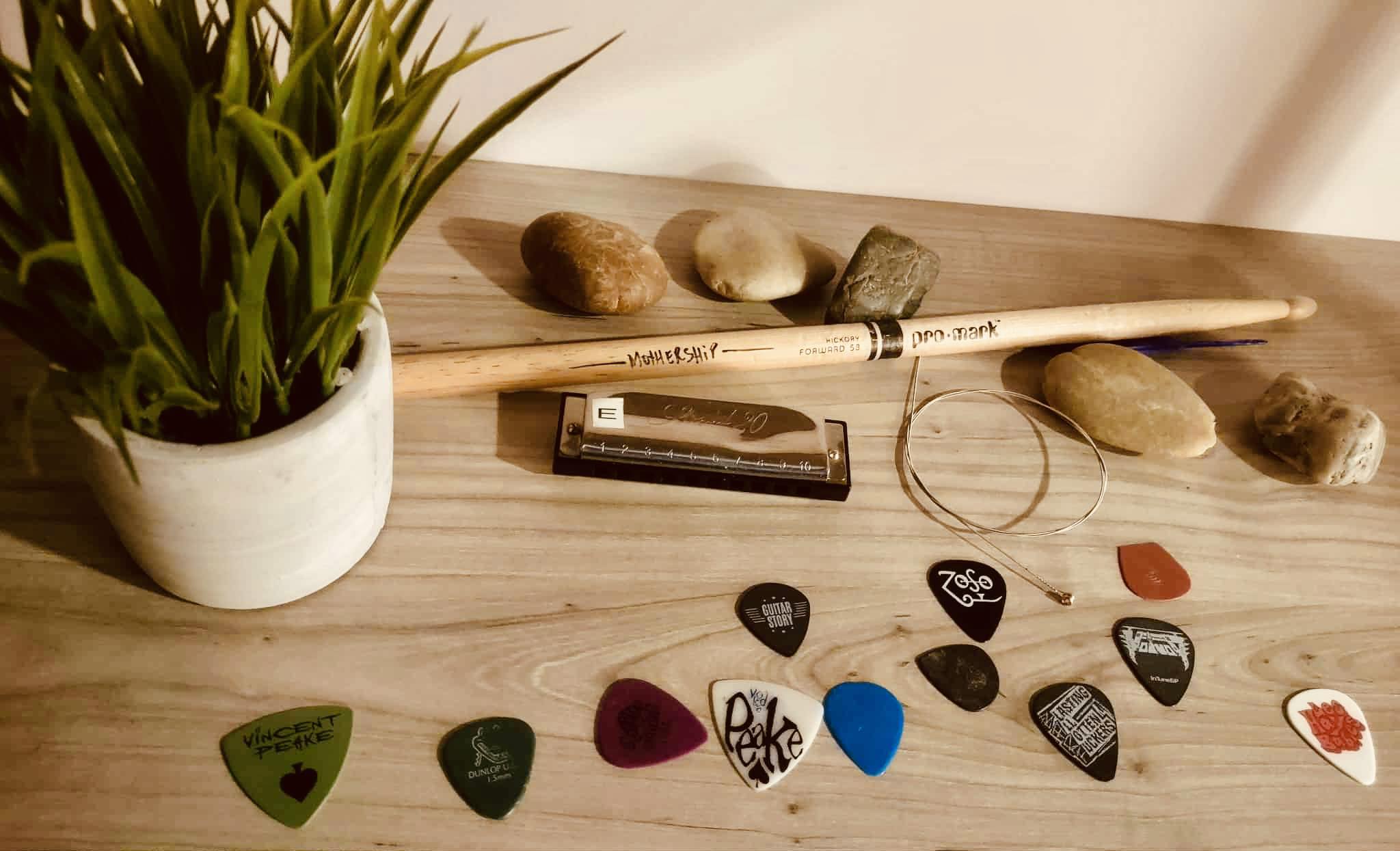 Une plante en pot, des pierres, deux baguettes, un harmonica, des médiators et des cordes de guitare sont disposés sur une surface en bois clair contre un mur uni.