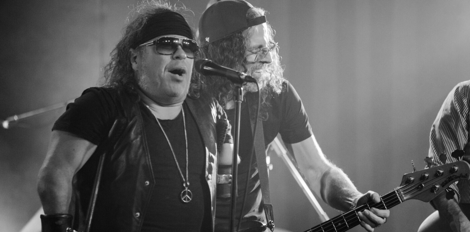 Deux musiciens se produisent sur scène ; l'un chante dans un microphone en portant des lunettes de soleil, un bandeau et un gilet, tandis que l'autre joue de la guitare basse et porte une casquette à l'envers. La scène est en noir et blanc.