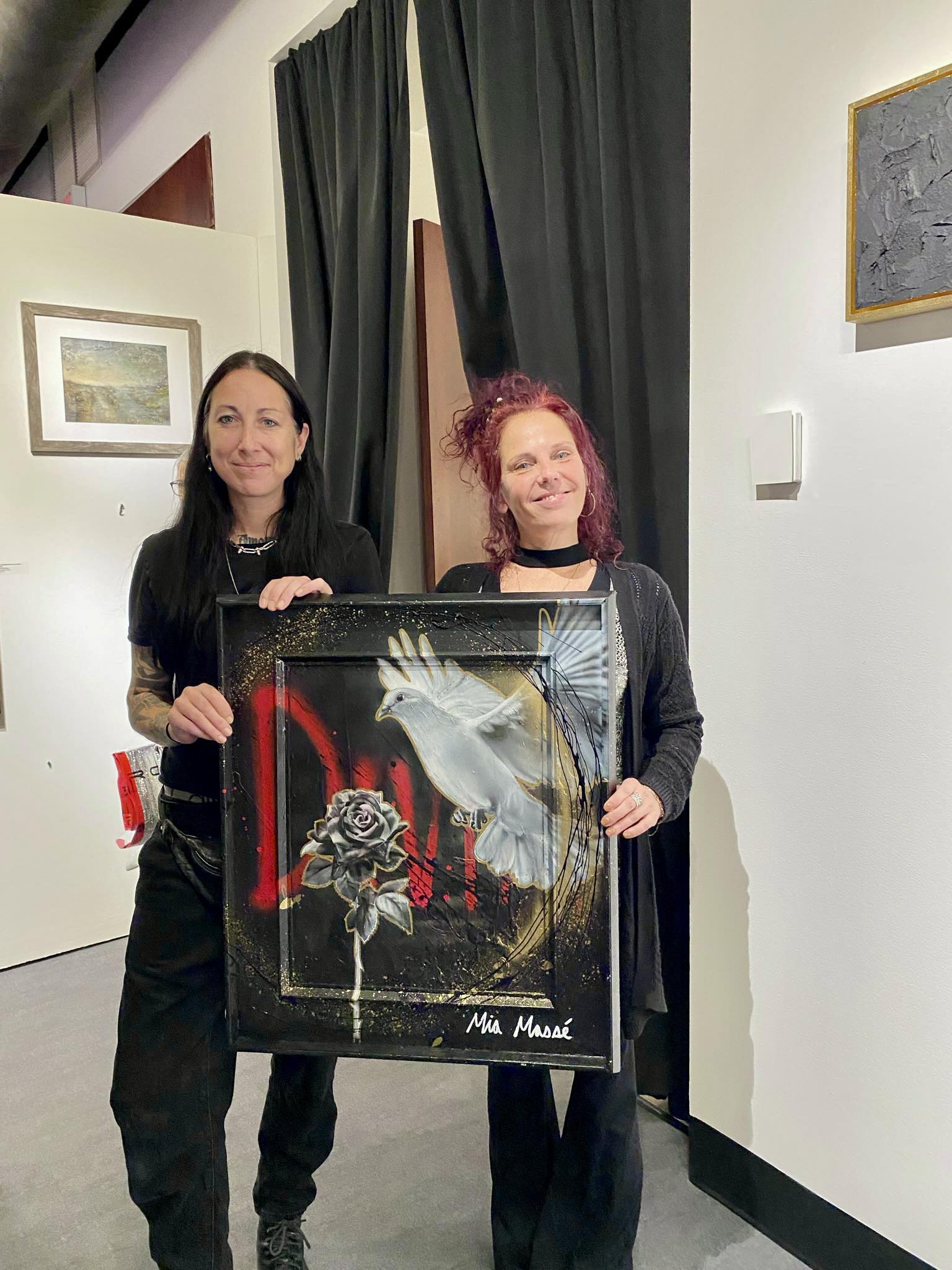 Deux femmes se tiennent à l'intérieur, souriantes et tenant un tableau représentant une colombe blanche, une rose noire et des touches audacieuses de rouge et d'or. Des œuvres d'art encadrées sont visibles sur les murs blancs derrière elles.