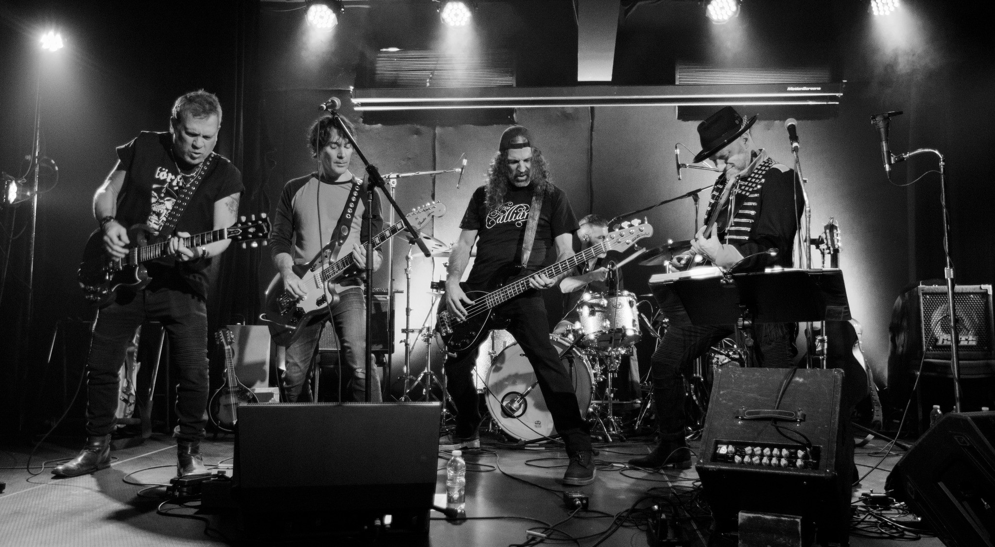 Photo en noir et blanc d'un groupe de rock composé de cinq membres, dont trois guitaristes, un bassiste et une personne jouant du clavier. Des lumières brillantes éclairent la scène et une batterie est visible à l'arrière-plan.