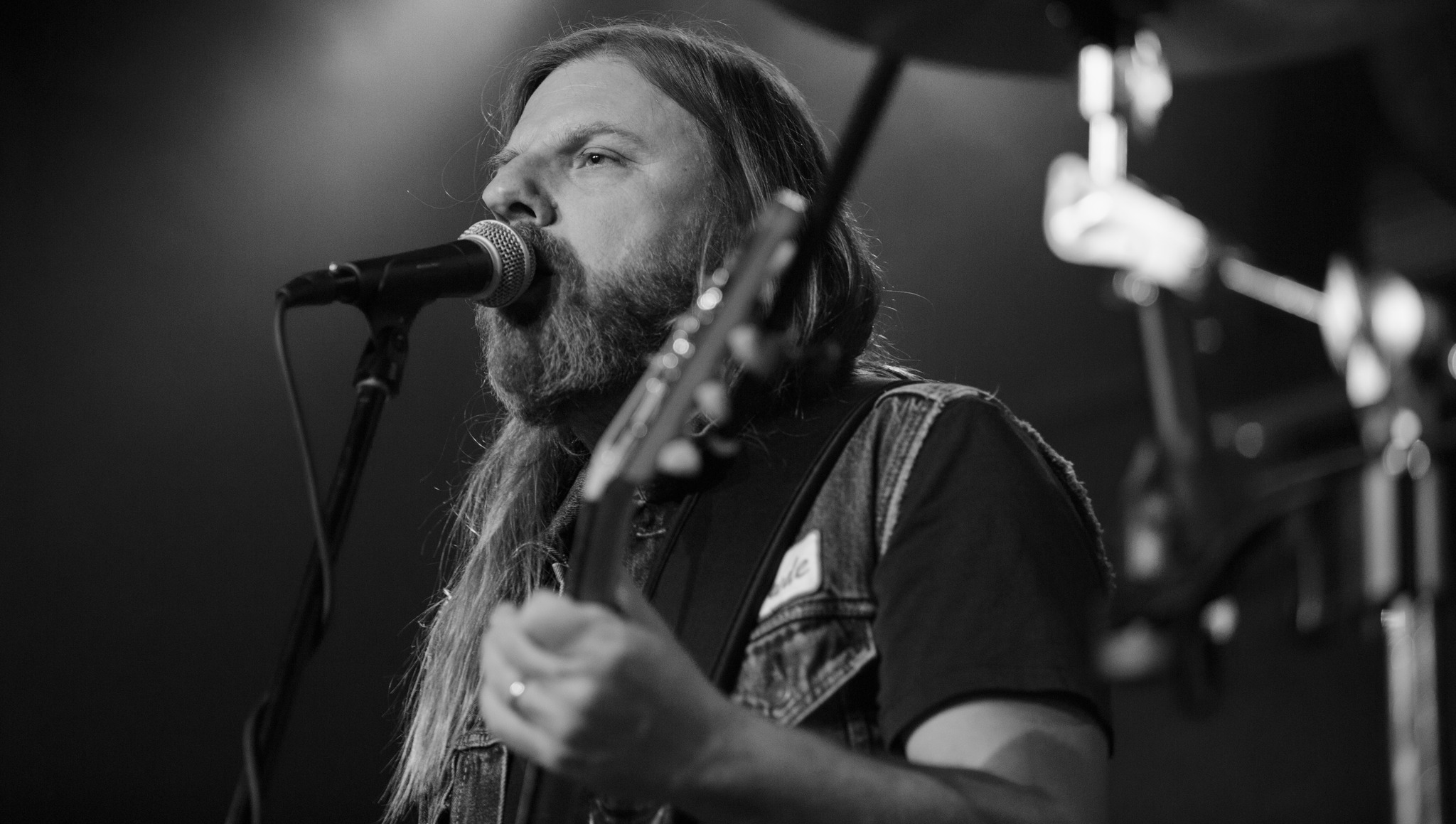Un musicien barbu aux cheveux longs chante dans un microphone tout en jouant de la guitare sur scène. L'image est en noir et blanc et l'arrière-plan est légèrement flou, ce qui permet de se concentrer sur l'artiste.