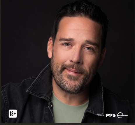 Un homme aux cheveux bruns courts et à la barbe, portant une veste en jean noire sur une chemise vert clair, sourit doucement sur un fond sombre uni. Des logos et une icône "18+" sont visibles dans les coins inférieurs.