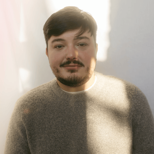Une personne aux cheveux courts et foncés, portant une moustache et une barbe, vêtue d'un pull-over gris, se tient debout dans une lumière naturelle douce avec des ombres sur un fond de couleur claire.